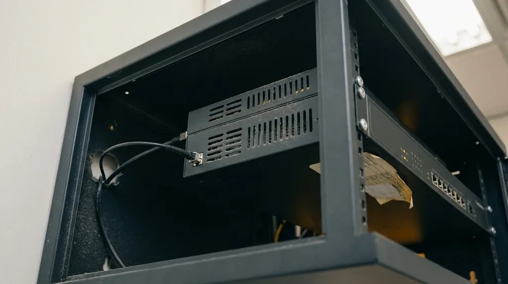 Router de perímetro montado en un armario de comunicaciones de pared, con un cable de fibra entrando por el lateral y una etiqueta de inventario despegada